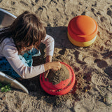 Ein Kind formt konzentriert Sand mit einer roten Kuppelform, während der Stapelstein: Gelb, ein zweiteiliger Form in Orange und Gelb, daneben liegt. Ein kleines Stück einer Rutsche ist sichtbar.