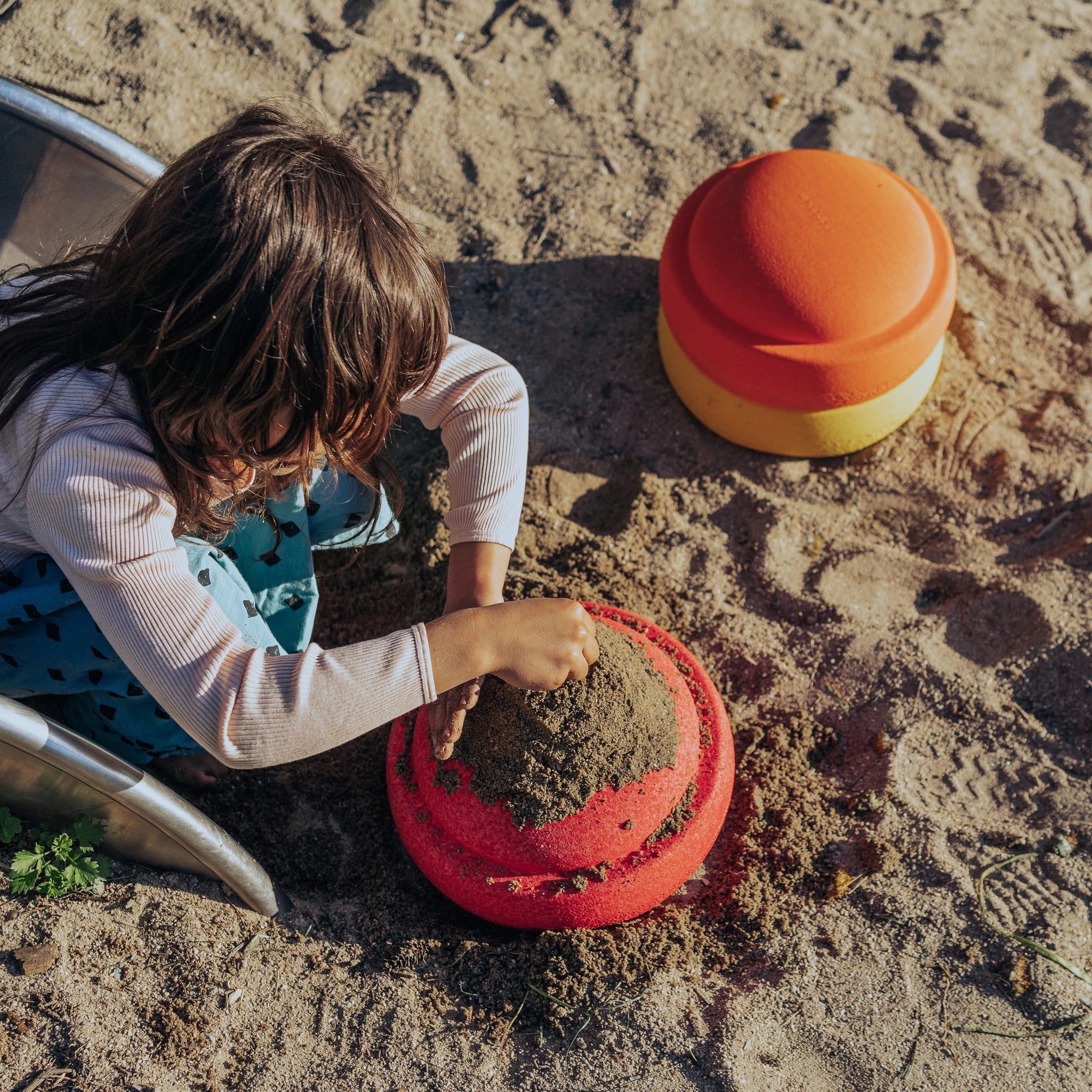 Ein Kind formt konzentriert Sand mit einer roten Kuppelform, während der Stapelstein: Gelb, ein zweiteiliger Form in Orange und Gelb, daneben liegt. Ein kleines Stück einer Rutsche ist sichtbar.