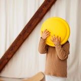 Kind steht drinnen mit Stapelstein: Regenbogen Classic (6 Steine), hält großen gelben Spielstein vor Gesicht. Im Hintergrund Holzbalken und gebogenes Balance-Board. Produkt verbindet Ästhetik und nachhaltiges Design von KLICKKLACK.