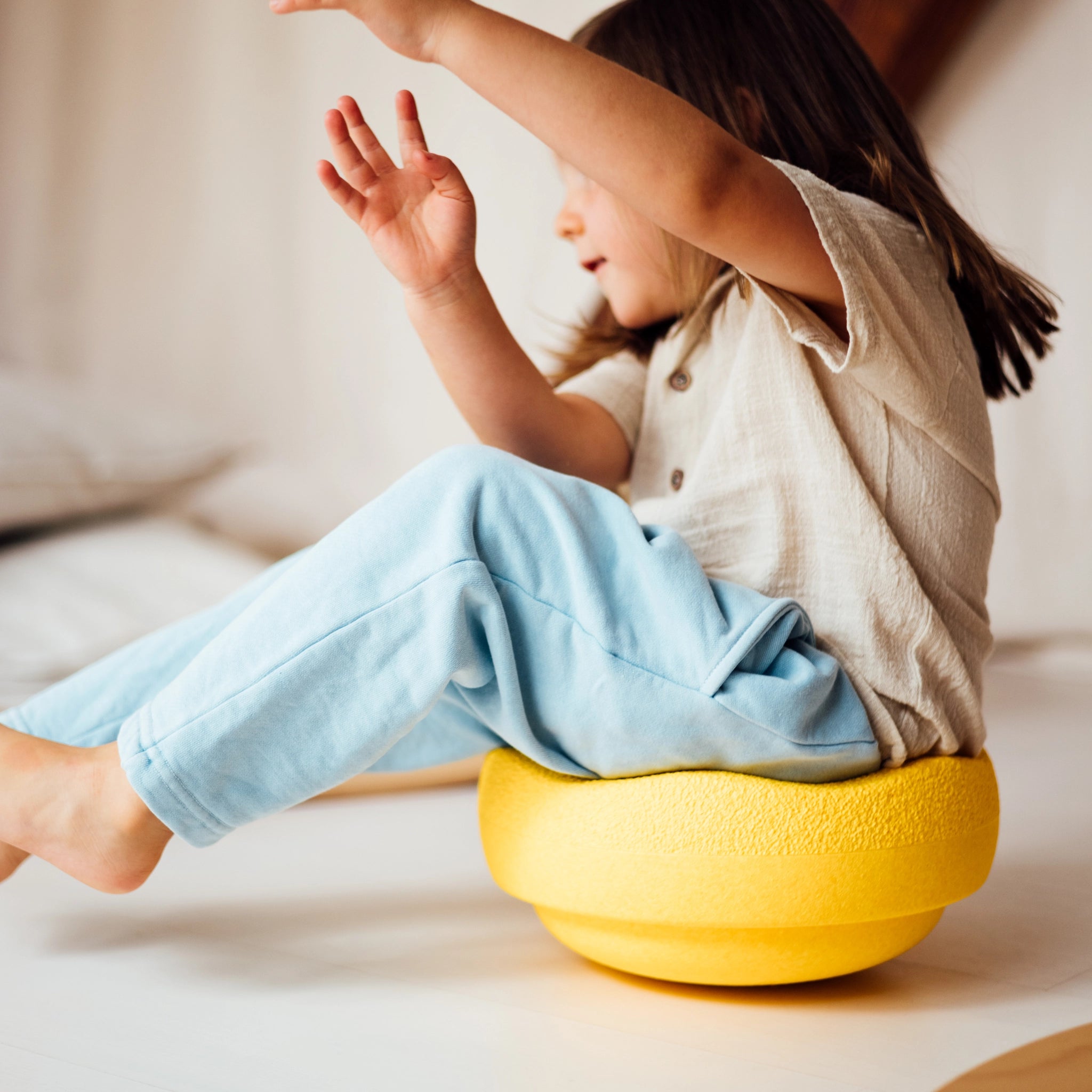 Kleinkind sitzt auf einem Stapelstein: Regenbogen Classic (6 Steine), einem runden, strukturierten gelben Kissen, und balanciert spielerisch. Die Szene vermittelt ein Gefühl von Leichtigkeit und Freude.