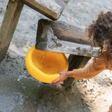 Ein Kind spielt in einem Sandbereich unter einer einfachen Holzspielstruktur. Es hält eine leuchtend gelbe Stapelstein-Pfanne, um den Sandfluss von einer kleinen Holzrampe aufzufangen.