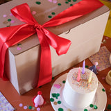 Geschenkbox stapelstein mit rotem Band auf Geburtstagstisch, umgeben von Konfetti, Kuchen und Partyhüten. Enthält Stapelstein: Regenbogen Classic (6+1 Balance Board), ideal für nachhaltige, moderne Feierlichkeiten von KLICKKLACK.