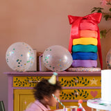 Ein Kindergeburtstags-Setup mit Regenbogenkissen „Stapelstein: Regenbogen Classic (6 Steine)“, dekorativ gestapelt und mit roter Schleife gebunden, auf lila Schrank neben Partyballons und Geschenkboxen.
