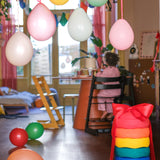 Stapelstein: Regenbogen Classic (6 Steine) im Vordergrund, dekoriert mit einer roten Schleife. Sie liegen in einem festlichen, kindgerechten Raum mit Luftballons und einem spielenden Kind am Tisch.