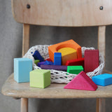 Bausteine: Bunte Geoklötze (30-teilig) aus hochwertigem Lindenholz in verschiedenen geometrischen Formen, präsentiert in einem Baumwollnetz auf einem Holzstuhl. Ideal für kreative Bauprojekte, handgefertigt in Deutschland.