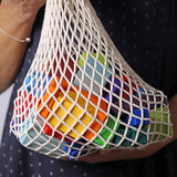 Bausteine: Bunte Geoklötze (30-teilig) in einem weißen Netz aus Baumwolle, gehalten von einer Hand mit silbernem Armband, zeigen bunte, geometrische Holzformen für kreatives Bauen, handgefertigt in Deutschland.