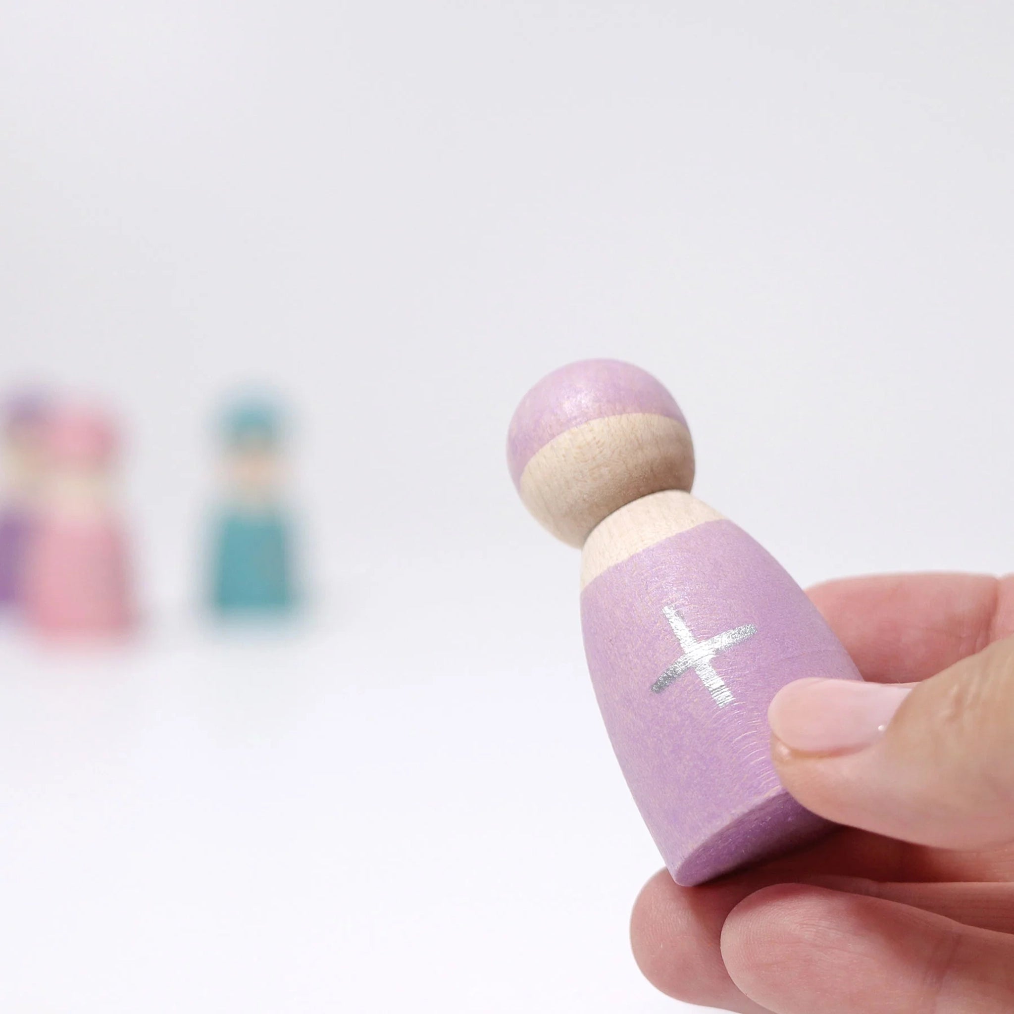 Freies Spiel: Fünf Rechenfreunde - Eine Hand hält eine lavendelfarbene, handbemalte Holzfigur mit natürlichem Kopf und einem + Zeichen. Ideal für haptisches Lernen und ein spielerischer Einstieg in Mathematik.