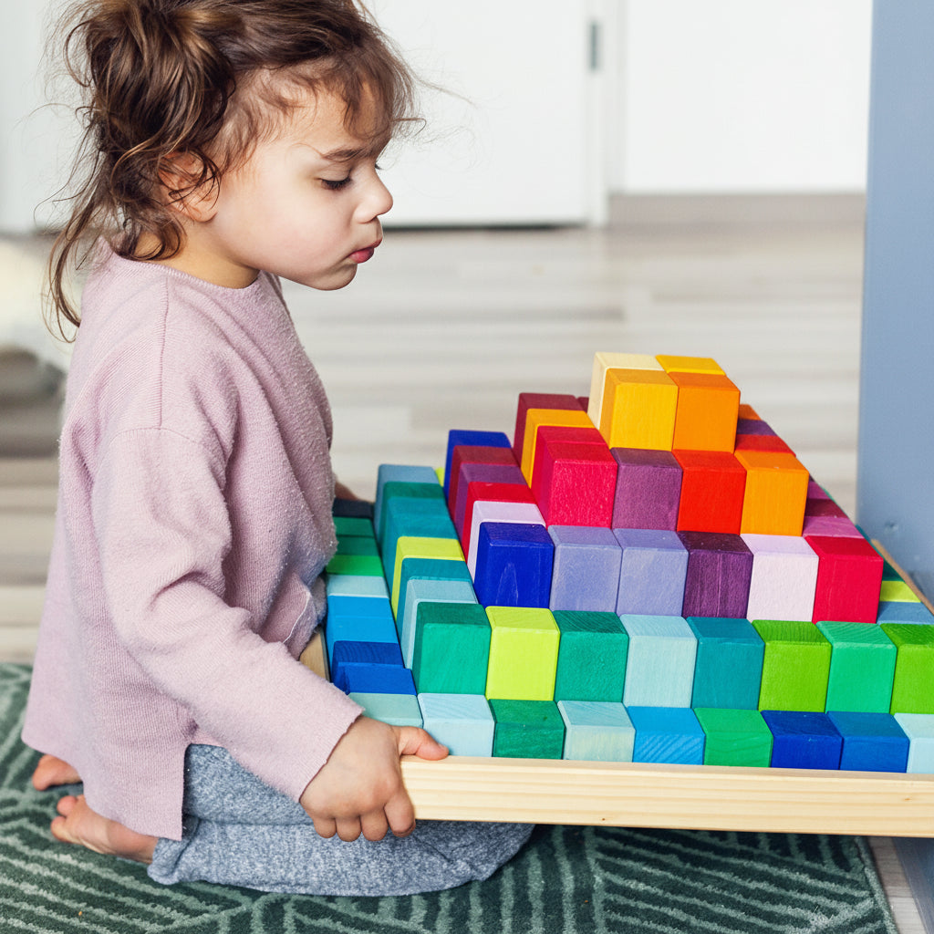 Ein Kleinkind sitzt auf einem Teppich und hält die Freies Spiel: Große Stufenpyramide, eine flache Holzschale mit kleinen, soliden Holz-Würfeln, die in einem stufenartigen, regenbogenähnlichen Muster angeordnet sind.