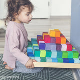 Ein Kleinkind sitzt auf einem Teppich und hält die Freies Spiel: Große Stufenpyramide, eine flache Holzschale mit kleinen, soliden Holz-Würfeln, die in einem stufenartigen, regenbogenähnlichen Muster angeordnet sind.