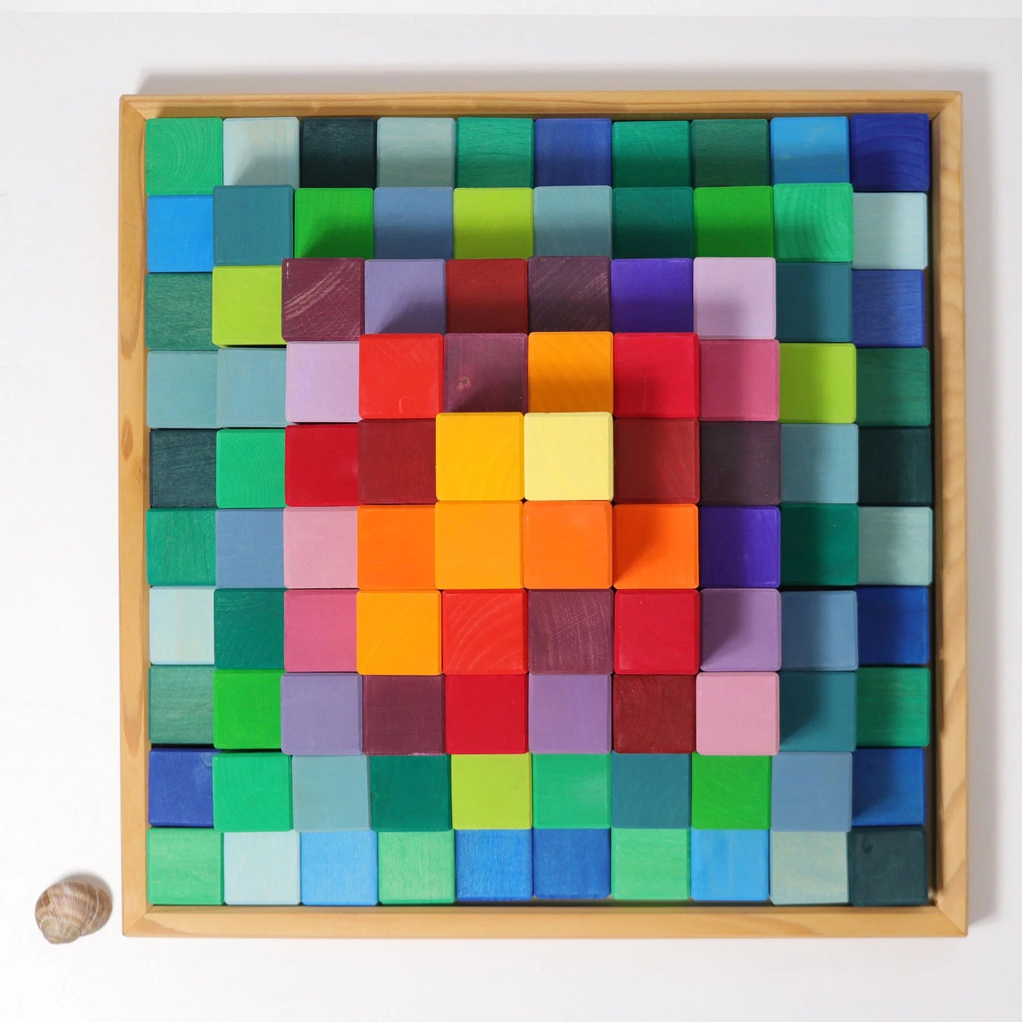 Freies Spiel: Große Stufenpyramide, eine flache Holzschale mit einem engmaschigen Raster aus kleinen, quadratischen Holzblöcken in Regenbogenfarben, die ein Farbverlaufsmuster bilden, daneben eine einzelne leere Schneckenschale.