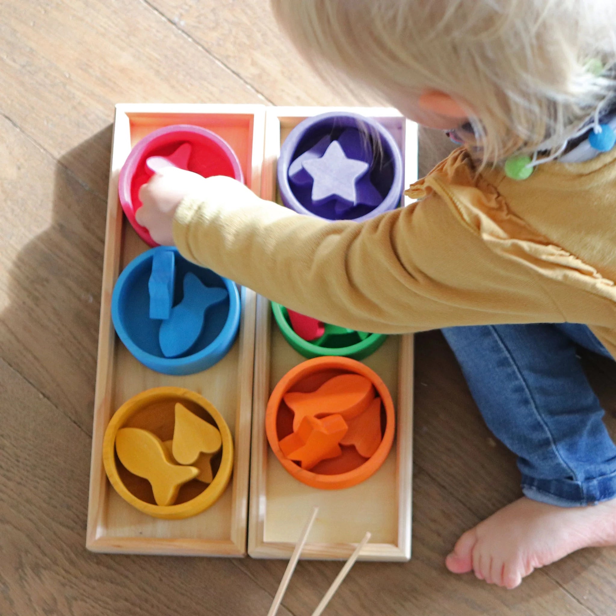 Freies Spiel: Sortierspiel Regenbogenschälchen aus geöltem Erlen- und lasiertem Lindenholz. Ein Kind sortiert bunte Holzformen mit einer Zange in zwei Holzrahmen mit sechs Schälchen. Fördert Feinmotorik und Konzentration.