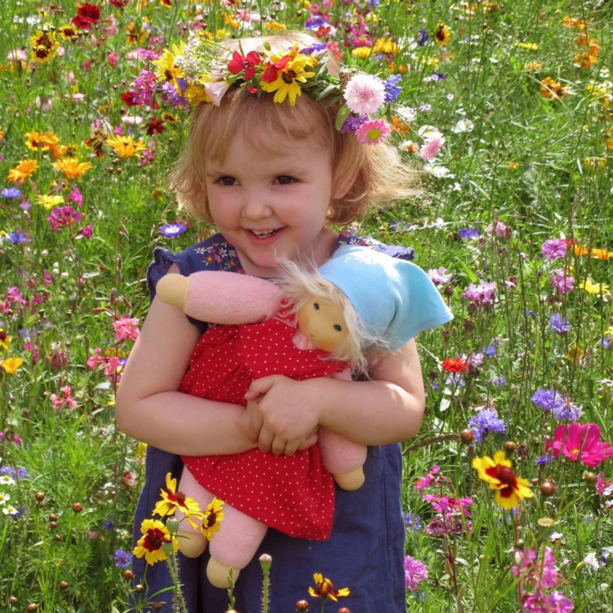 Mädchen in Blumenwiese umarmt Puppe: Sommerkind Lotti, aus Bio-Baumwollfrottee und schadstofffreier Schafschurwolle, mit an- und ausziehbarem Kleid. Handgefertigt in Deutschland, symbolisiert nachhaltige KLICKKLACK-Ästhetik.