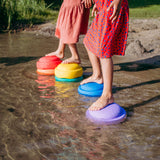 Zwei Kinder balancieren über das flache Wasser auf dem Stapelstein: Regenbogen Classic (6+1 Balance Board), bestehend aus bunten, kuppelförmigen Schaumstoff-Steinen, die spielerisch eine Regenbogenfolge bilden.