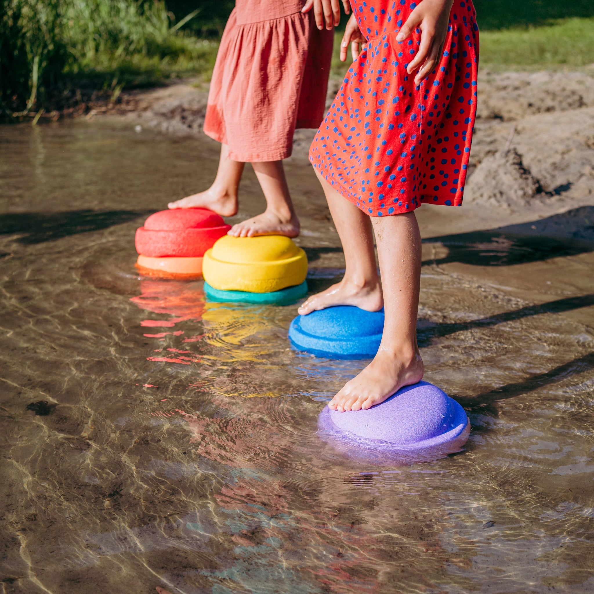 Zwei Kinder balancieren über das flache Wasser auf dem Stapelstein: Regenbogen Classic (6+1 Balance Board), bestehend aus bunten, kuppelförmigen Schaumstoff-Steinen, die spielerisch eine Regenbogenfolge bilden.