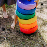 Ein Kind stapelt im Freien sechs Stapelstein: Regenbogen Classic (6 Steine) in Form von großen, donutartigen Schaumstoffringen auf Gras und Erde. Die Ringe sind harmonisch angeordnet und fördern spielerische Kreativität.