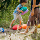 Zwei Kinder spielen auf einer Wiese mit Stapelstein: Regenbogen Classic (6 Steine), farbenfrohe Balance-Discs. Ein Kind trägt zwei Steine, während das andere über die leuchtenden Schaumstoffpads balanciert.