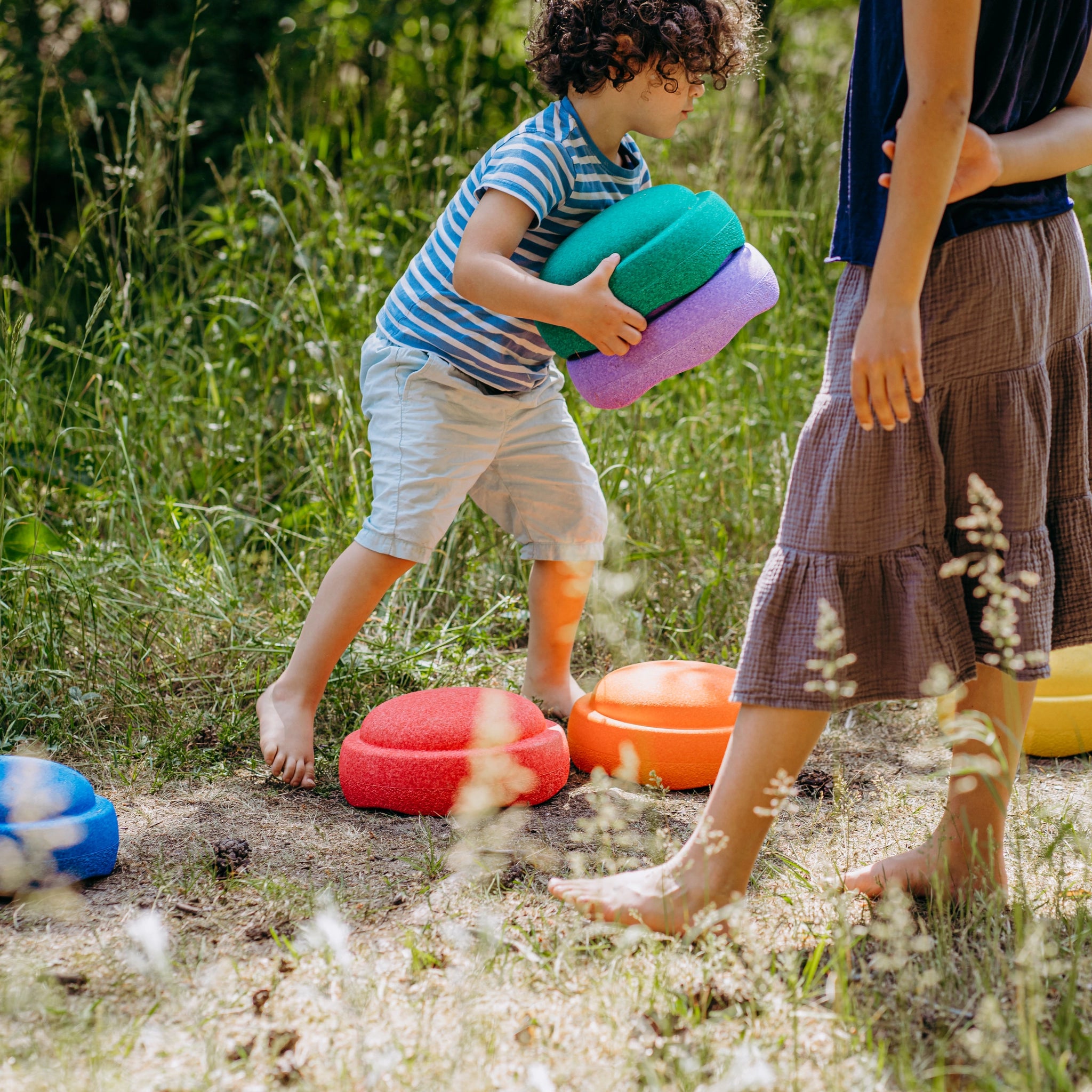 Zwei Kinder spielen auf einer Wiese mit Stapelstein: Regenbogen Classic (6 Steine), farbenfrohe Balance-Discs. Ein Kind trägt zwei Steine, während das andere über die leuchtenden Schaumstoffpads balanciert.