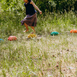 Ein Kind springt barfuß über die Stapelstein: Regenbogen Classic (6 Steine) in einem sonnigen Garten. Die leuchtenden Steine bilden einen kreativen Parcours auf dem grasbewachsenen Boden.