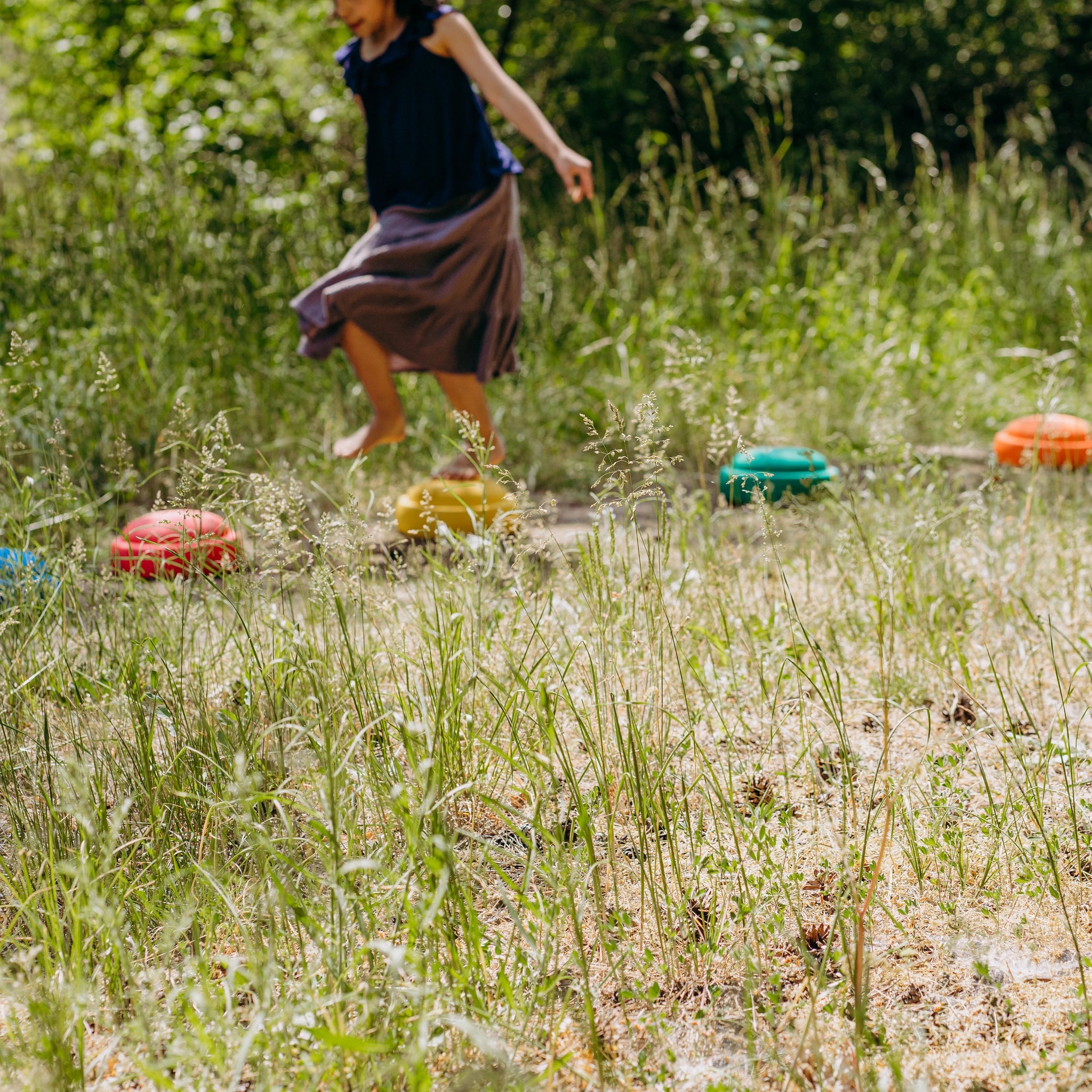 Ein Kind springt barfuß über die Stapelstein: Regenbogen Classic (6 Steine) in einem sonnigen Garten. Die leuchtenden Steine bilden einen kreativen Parcours auf dem grasbewachsenen Boden.