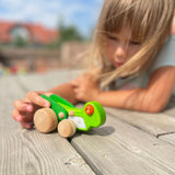 Kind spielt mit dem Rückziehspielzeug: Grashüpfer auf einer Holzterrasse. Das grüne Holzspielzeug hebt beim Zurückziehen seine Flügel. Hergestellt aus Buchenholz in einer polnischen Manufaktur.